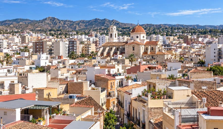 Tiled rooftops and pale apartment blocks climb towards a stone church with a terracotta dome, under a bare brown mountain ridge and a clear blue sky